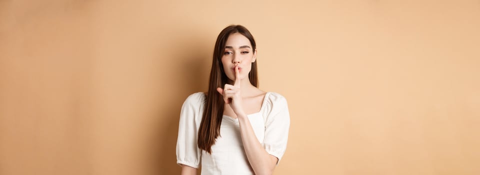 Sensual cute girl hiding secret, showing hush gesture, make shhh sound with finger on lips, stay quiet sign, standing on beige background photo