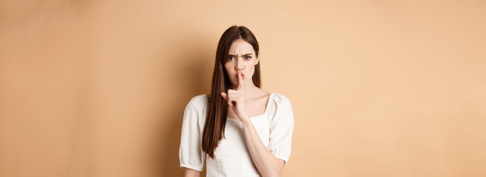 Angry young girl scolding bad behavious, shushing and frowning mad, tell to be quiet, looking displeased, standing on beige background photo