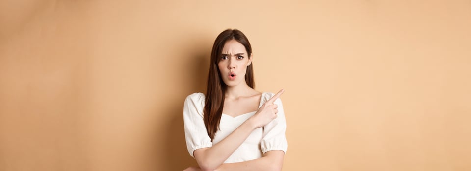 What is that. Shocked and angry woman scolding person, frowning and pointing left with confused face, waiting for explanation, standing on beige background photo