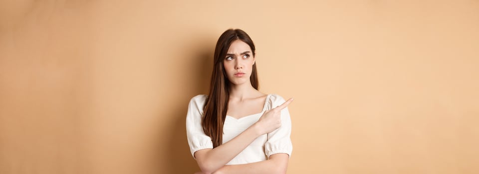 Suspicious woman in dress looking, pointing left, frowning and staring with disbelief and doubts, standing on beige background photo