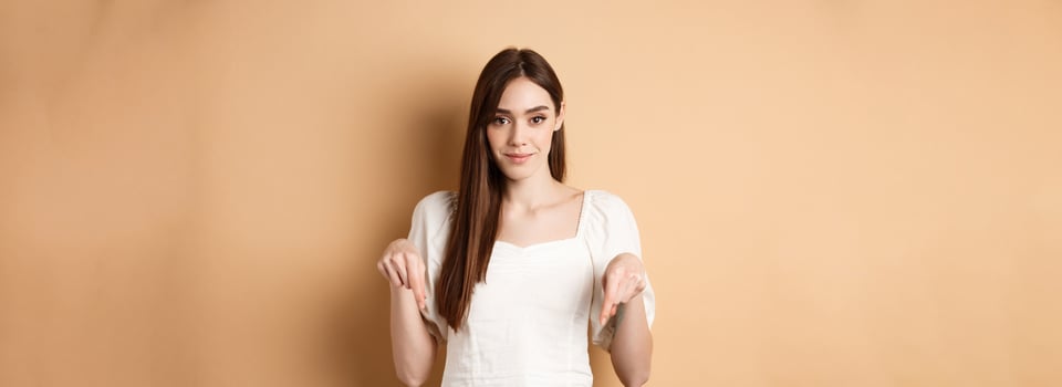 Smiling tender girl in dress pointing fingers down, showing advertisement on empty space, beige background photo