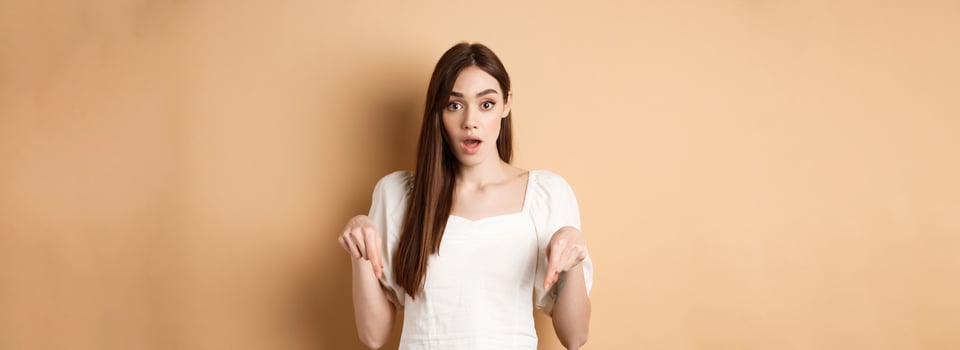 Wow look there. Amazed young woman in white dress pointing fingers down, standing in awe with dropped jaw and popped eyes against beige background photo