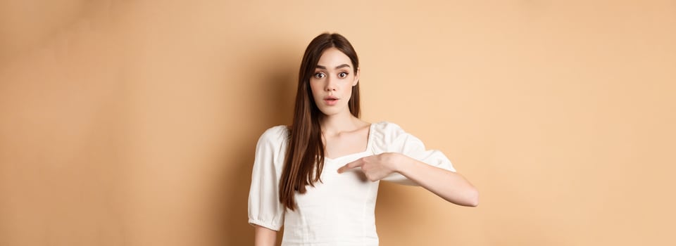 Confused girl pointing at herself with disbelief, being chosen or picked, standing on beige background photo