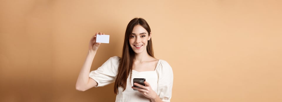 E-commerce concept. Smiling woman showing plastic credit card while shopping online on mobile phone, standing against beige background photo