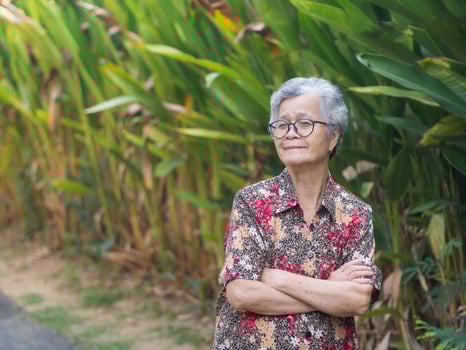 Cheerful senior woman with short gray hair, wearing glasses, smiling, arms crossed and looking at the camera while standing in a garden. Concept of aged people and relaxation photo