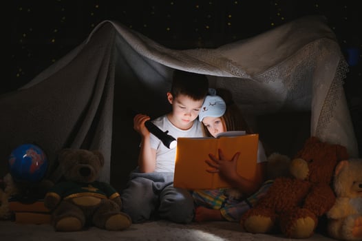 Two children with flashlight read a book under a blanket as a tent photo