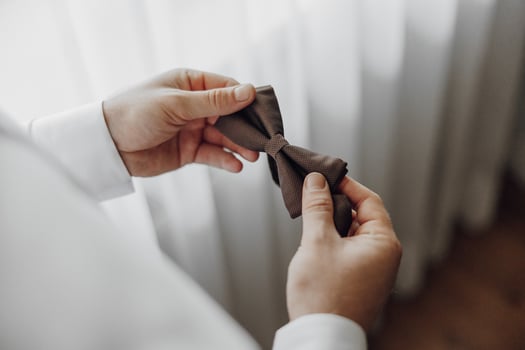 A man is holding a tie in his hand. The tie is brown and has a plaid pattern. The man is wearing a white shirt and he is getting ready for a formal event photo