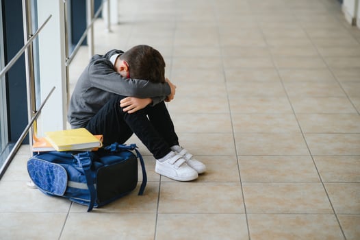 Little boy sitting alone on floor after suffering an act of bullying while children run in the background. Sad young schoolboy sitting on corridor with hands on knees and head between his legs photo
