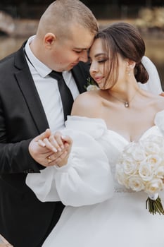 A bride and groom are embracing each other in a wedding photo. The bride is wearing a white dress and the groom is wearing a black suit photo