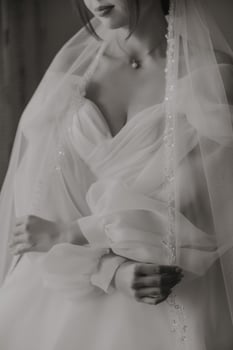 A woman in a white dress is holding a veil in her hand. The dress is white and the veil is made of a material that looks like lace. The woman is getting ready for a special occasion photo
