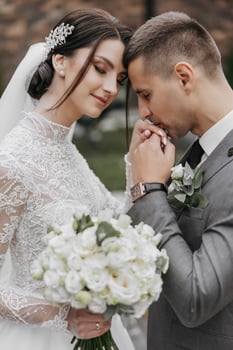 A bride and groom are embracing each other with their hands on their faces. The bride is holding a bouquet of flowers and the groom is wearing a watch. Scene is one of love and happiness photo