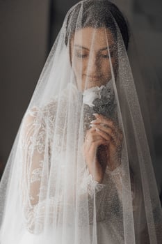 A bride is wearing a veil and holding a flower. The veil is made of a net-like material and covers her face photo