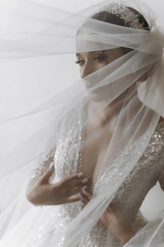 A woman is wearing a white veil and a white dress. She is standing in front of a white wall photo