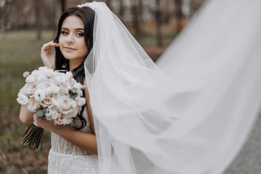 A bride is holding a bouquet of flowers and standing in front of a white veil. The scene is set in a field, with trees in the background. The bride's dress is white, and she has a smile on her face photo