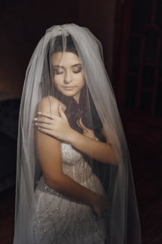 A woman in a wedding dress is wearing a veil. She is looking at the camera. The image has a romantic and intimate mood photo