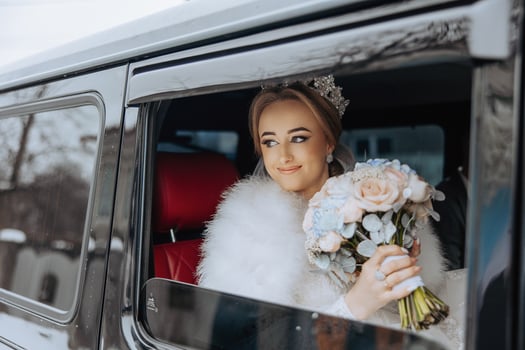 A bride is sitting in a limousine with a bouquet of flowers. She is wearing a white fur coat and a tiara photo
