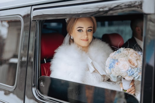 A bride is sitting in a car with a bouquet of flowers. She is wearing a white fur coat and a tiara photo