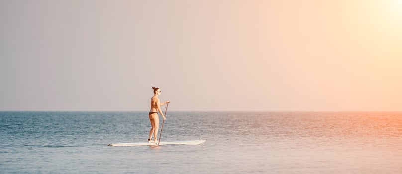 Sea woman sup. Silhouette of happy middle aged woman in rainbow bikini, surfing on SUP board, confident paddling through water surface. Idyllic sunset. Active lifestyle at sea or river. photo