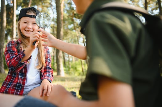 Group of curious happy school kids in casual clothes with backpacks exploring nature and forest together on sunny autumn day, girl holding magnifying glass and looking at fir cone in boy hands. photo