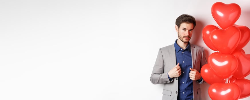 Lovers day. Handsome and confident young man getting dressed for Valentines day, fixing suit and looking at camera, standing near romantic heart balloons, white background photo