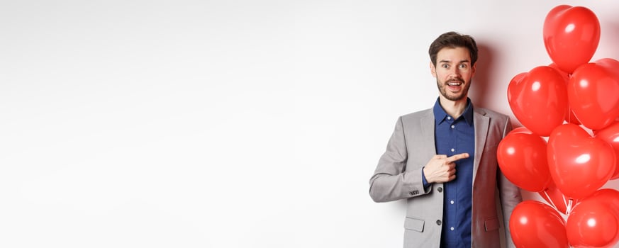 Lovers day. Excited male model in suit pointing finger at Valentines heart balloons and smiling, prepare romantic gifts on date, standing over white background photo