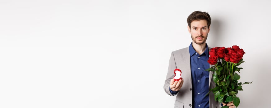 Handsome man in suit going to make a proposal, standing with red roses flowers and engagement rin in box, making romantic surprise, white background photo