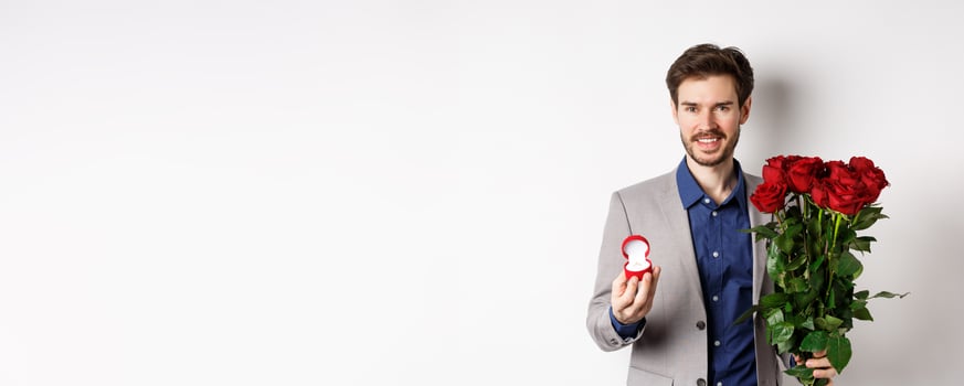 Smiling boyfriend making a marriage proposal, standing with engagement ring and bouquet of red roses, going on romantic Valentines day date, white background photo