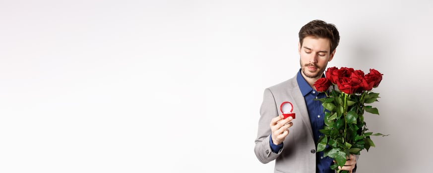 Romantic man in suit looking pensive at engagement ring, going to make a marriage proposal on Valentines day, holding bouquet of roses, standing over white background photo
