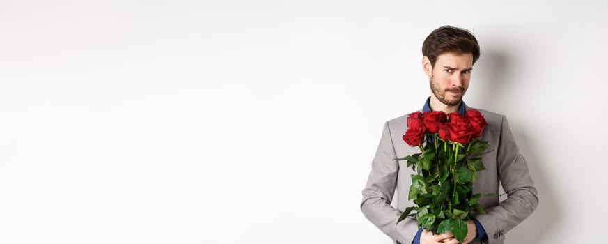 Indecisive bearded man in suit, holding flowers roses and looking at camera unsure, going on romantic date, standing over white background photo
