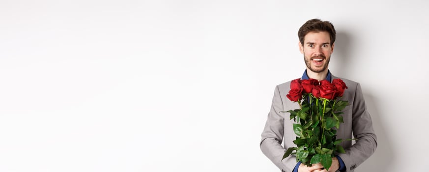 Excited handsome man in suit holding bouquet of roses for romantic date with lover, standing happy on Valentines day, white background photo