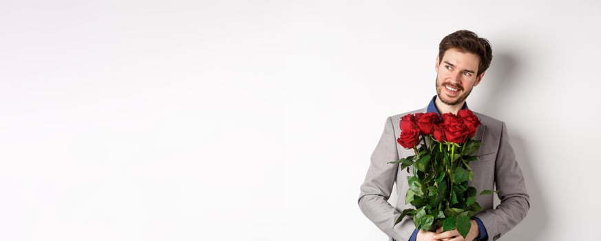 Charming smiling man going on romantic date, wearing suit, holding bouquet of roses and looking left, standing on valentines day with gift on white background photo