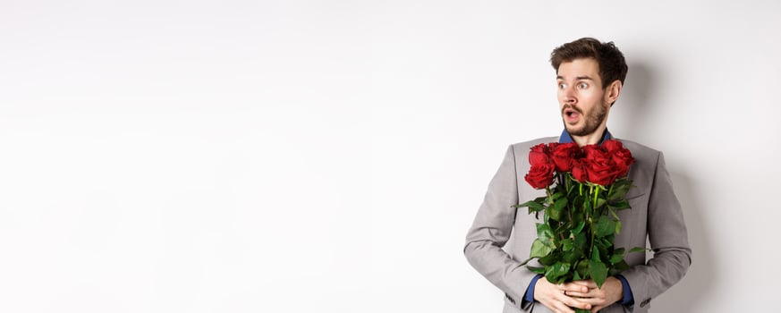 Handsome young man in suit holding red roses, looking left with surprised and startled expression, standing on Valentines day over white background photo