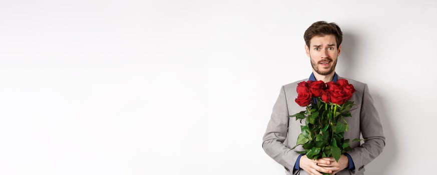 Worried boyfriend in suit, holding flowers roses and looking doubtful at camera, standing with bouquet on valentines day against white background photo