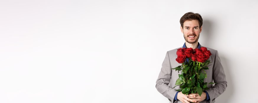 Handsome young man in suit standing with gift on valentines day, holding red roses bouquet and smiling at lover, standing over white background photo