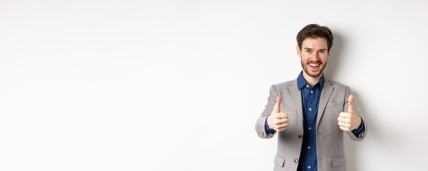 Cheerful and positive young man in business suit showing thumbs up and smiling, like something good, praise good job, standing happy on white background photo
