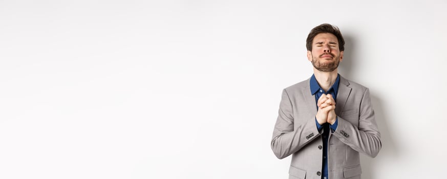 Nervous male entrepreneur in suit begging god, holding hands in pray with pleading expression, close eyes and make wish, asking for help, standing on white background photo
