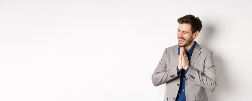 Happy and thankful businessman rejoicing, holding hands in begging or namaste sign, thanking for help, smiling relieved, standing in suit on white background photo