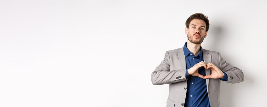 Romantic man in suit showing heart sign and pucker lips for kiss, express love and passion, like his lover, standing on white background photo