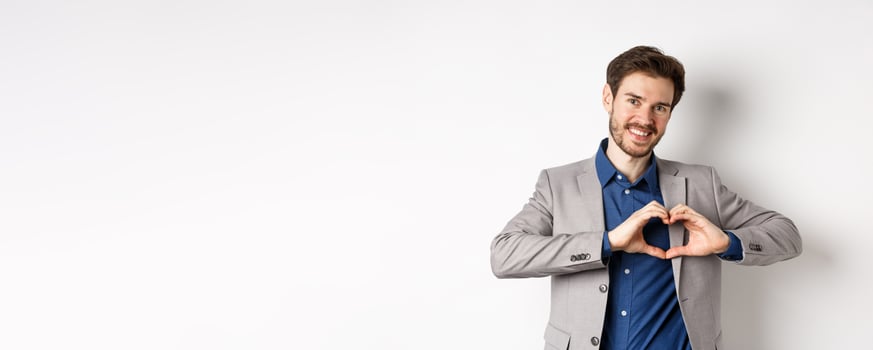 Romantic man in suit showing heart sign and smiling, love his girlfriend, standing on white background, making confession to lover photo