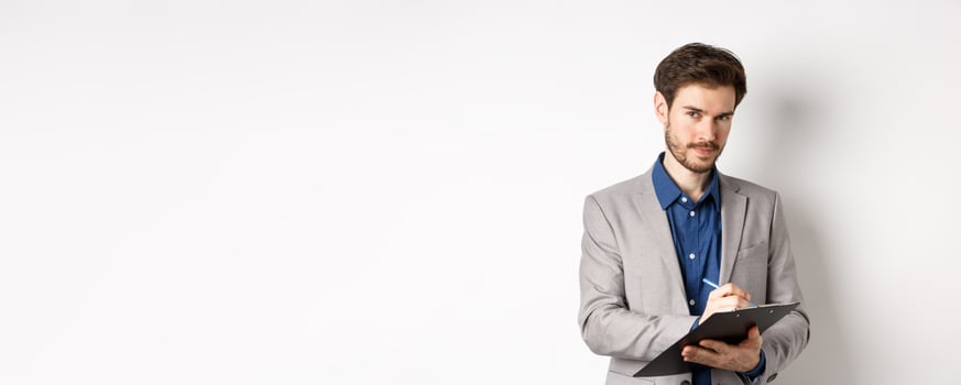 Successful businessman in grey suit taking notes, working in office clothing, holding clipboard and looking at camera, white background photo