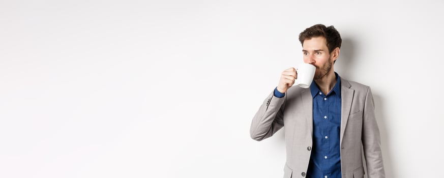 Handsome businessman in suit drinking coffee or tea from office mug, looking aside at logo, standing against white background photo