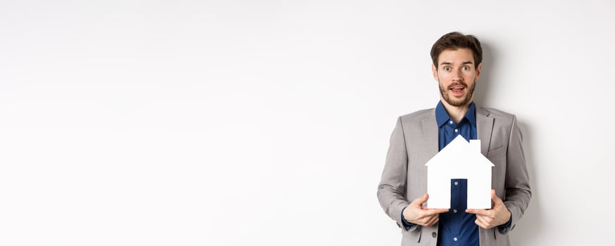 Real estate. Excited business man in suit holding paper house cutout and looking amazed at camera, buying apartment, white background photo