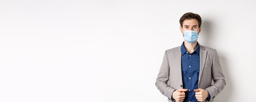 Covid-19 and office workers concept. Confident businessman in medical mask and suit looking at camera, standing on white background photo