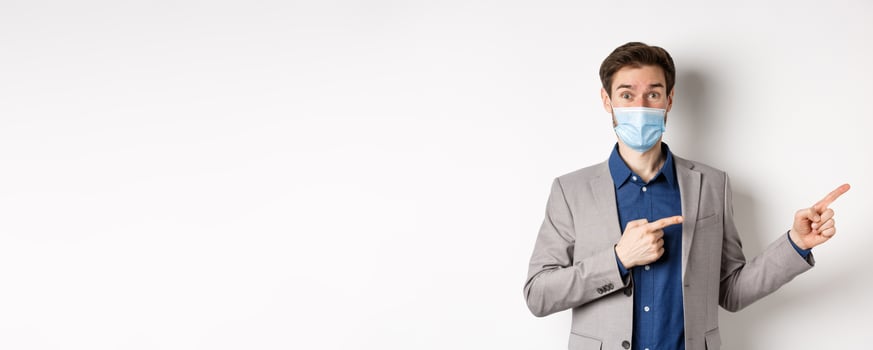 Covid-19, pandemic and business concept. Excited male entrepreneur in medical mask and suit pointing fingers right and looking curious, white background photo