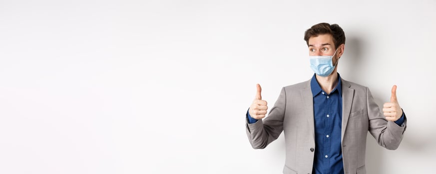 Covid-19, pandemic and business concept. Excited businessman in face mask and suit, showing thumbs up and looking aside at logo impressed, praise good thing, white background photo
