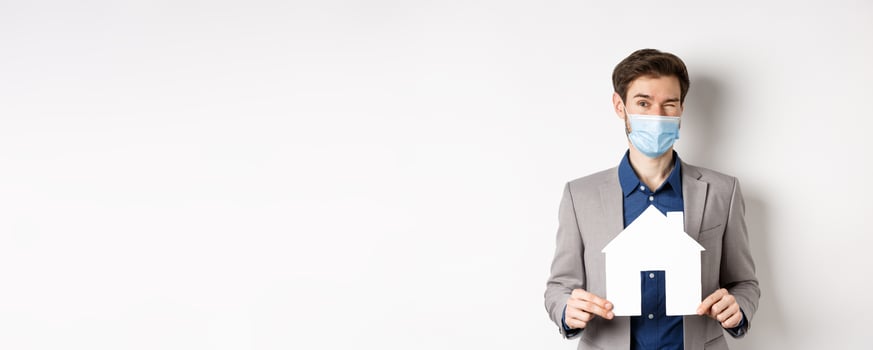Real estate and covid-19 concept. Cheerful young man in medical mask and suit winking at camera, advertising new flats, showing paper house cutout photo