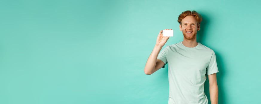 Shopping concept. Handsome redhead man in t-shirt showing plastic credit card and smiling, standing over turquoise background photo