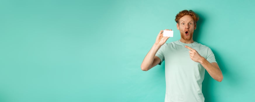 Shopping concept. Handsome redhead man in t-shirt showing plastic credit card and smiling, standing over turquoise background photo