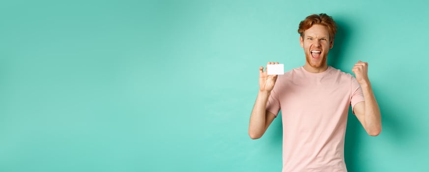 Cheerful young man triumphing, making fist pump to celebrate success, showing plastic credit card, winning prize from bank, turquoise background photo