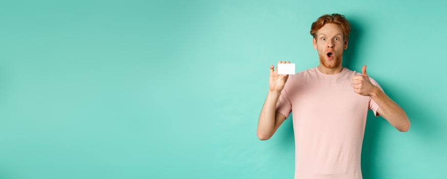 Adult guy with red hair and beard showing plastic credit card and thumb up, looking impressed, recommend bank, standing over mint background photo